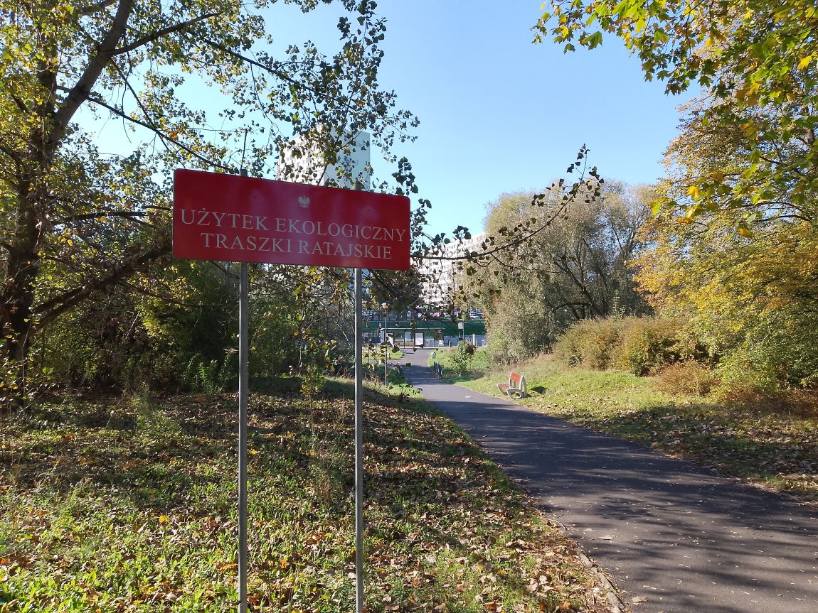 Tablica z napisem UŻYTEK EKOLOGICZNY TRASZKI RATAJSKIE wokół zieleń drzewa trawa, ścieżka asfaltowa, ławka, w oddali wiata przystankowa - grafika artykułu