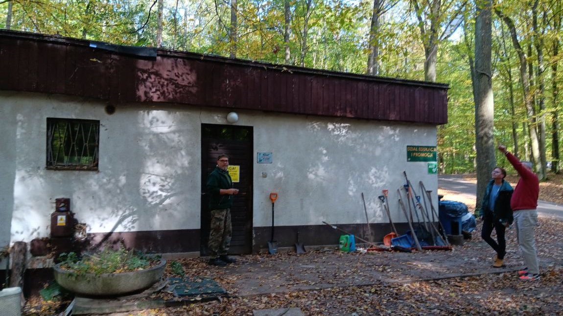 Niski nieduży budynek parterowy, w otoczeniu drzew, przed budynkiem trzy osoby