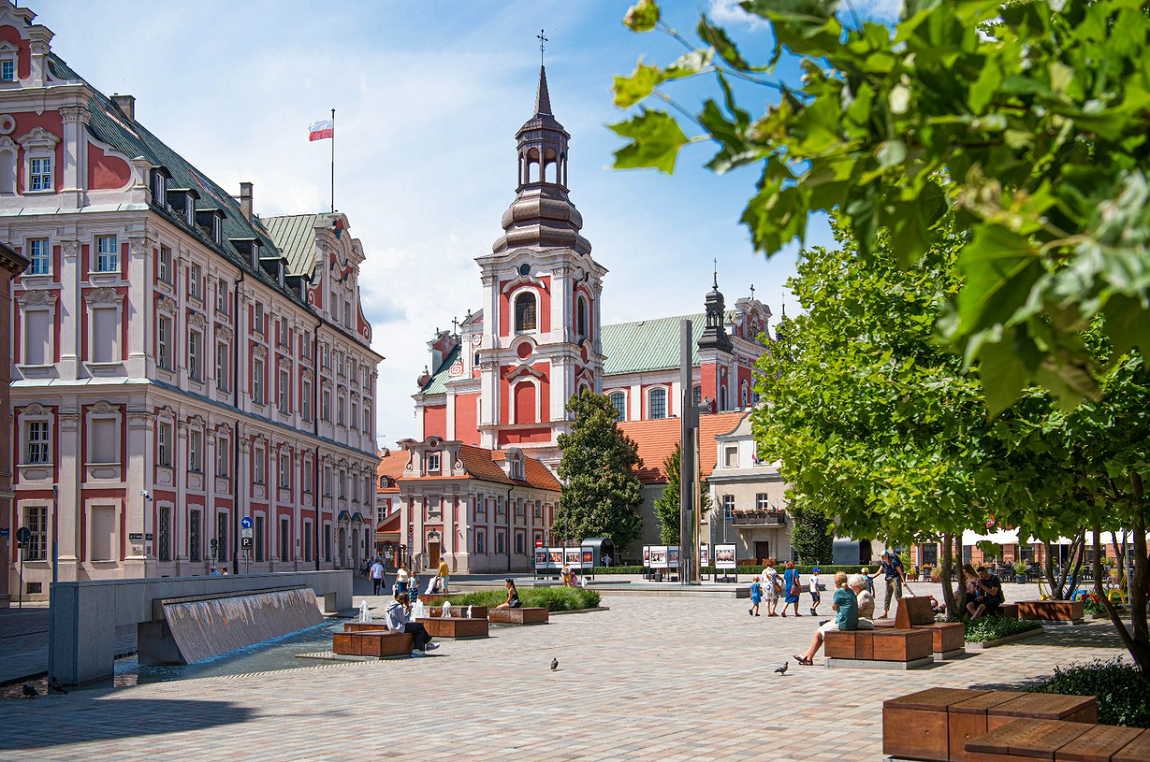 Widok z placu Kolegiackiego na Urząd Miasta Poznania, na placu spracerują ludzie lub siedzą na ławkach, na gmachu Urzędu powiewa biało czerwona flaga - grafika artykułu