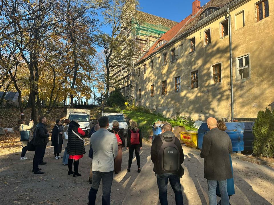 gruoa osób stoi przed wysokim budynkiem, obok drzewo, świcie słońce - grafika artykułu