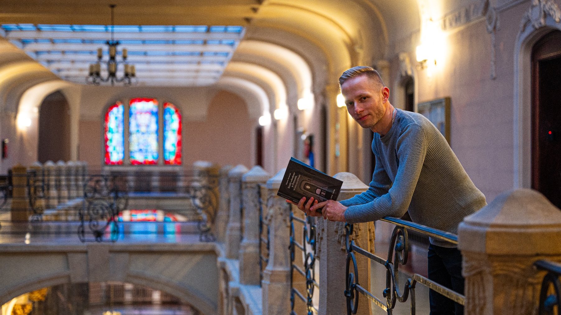 Na zdjęciu bogato zdobiona klatka schodowa, w centrum mężczyzna trzymający książkę