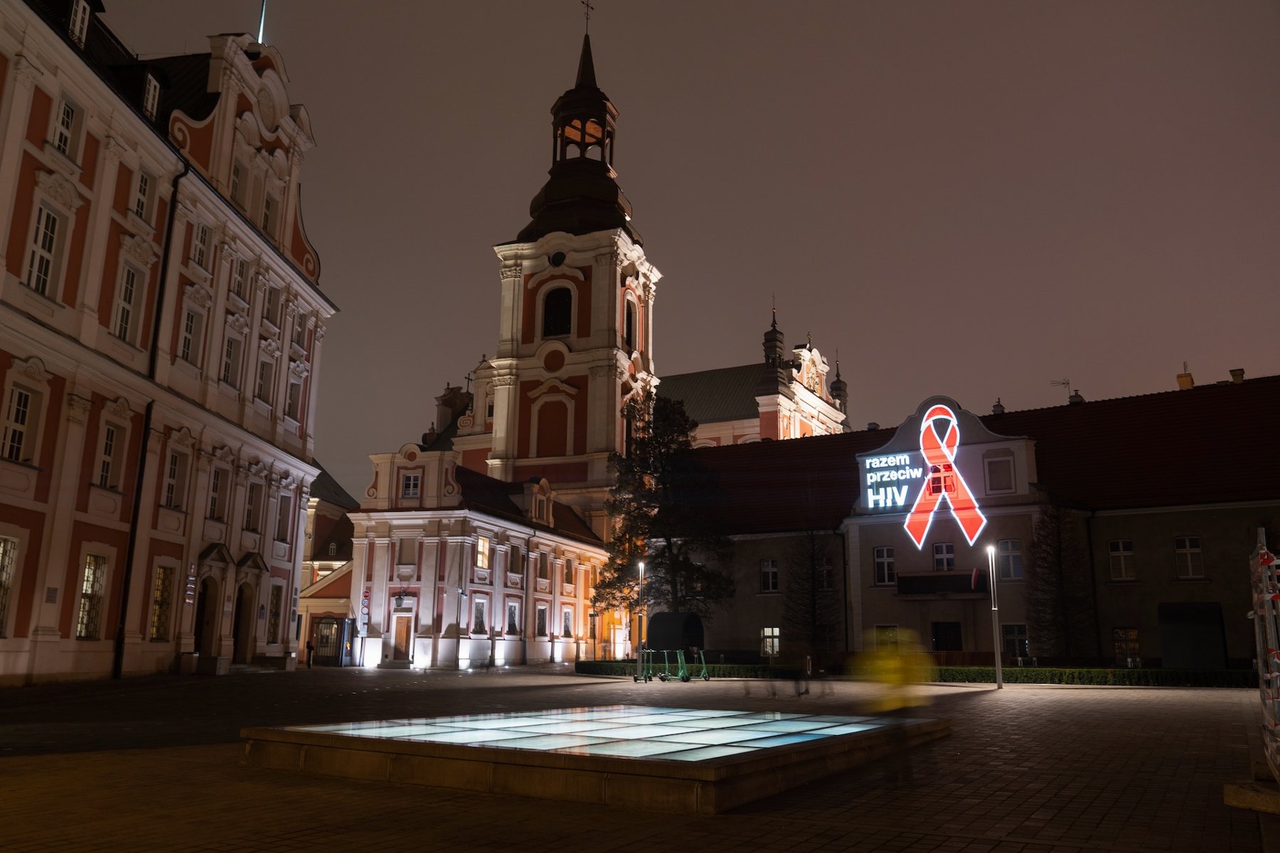 Zdjęcie przedstawia Urząd Miasta Poznania i budynki obok, na których znajduje się podświetlona kokarda i napis razem przeciwko HIV. - grafika artykułu