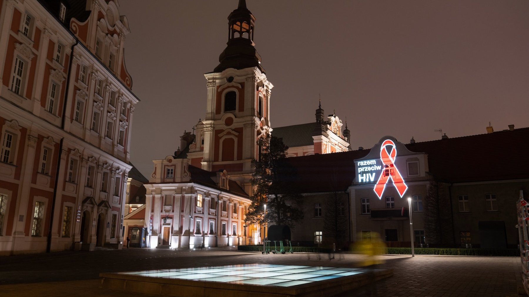 Zdjęcie przedstawia Urząd Miasta Poznania i budynki obok, na których znajduje się podświetlona kokarda i napis razem przeciwko HIV.