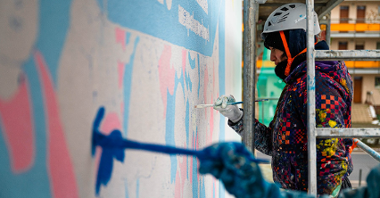 Na zdjęciu dwie kobiety na rusztowaniu, malujące mural