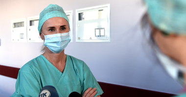 Na zdjęciu lekarka mówiąca coś do mikrofonu, stojąca w szpitalnym korytarzu