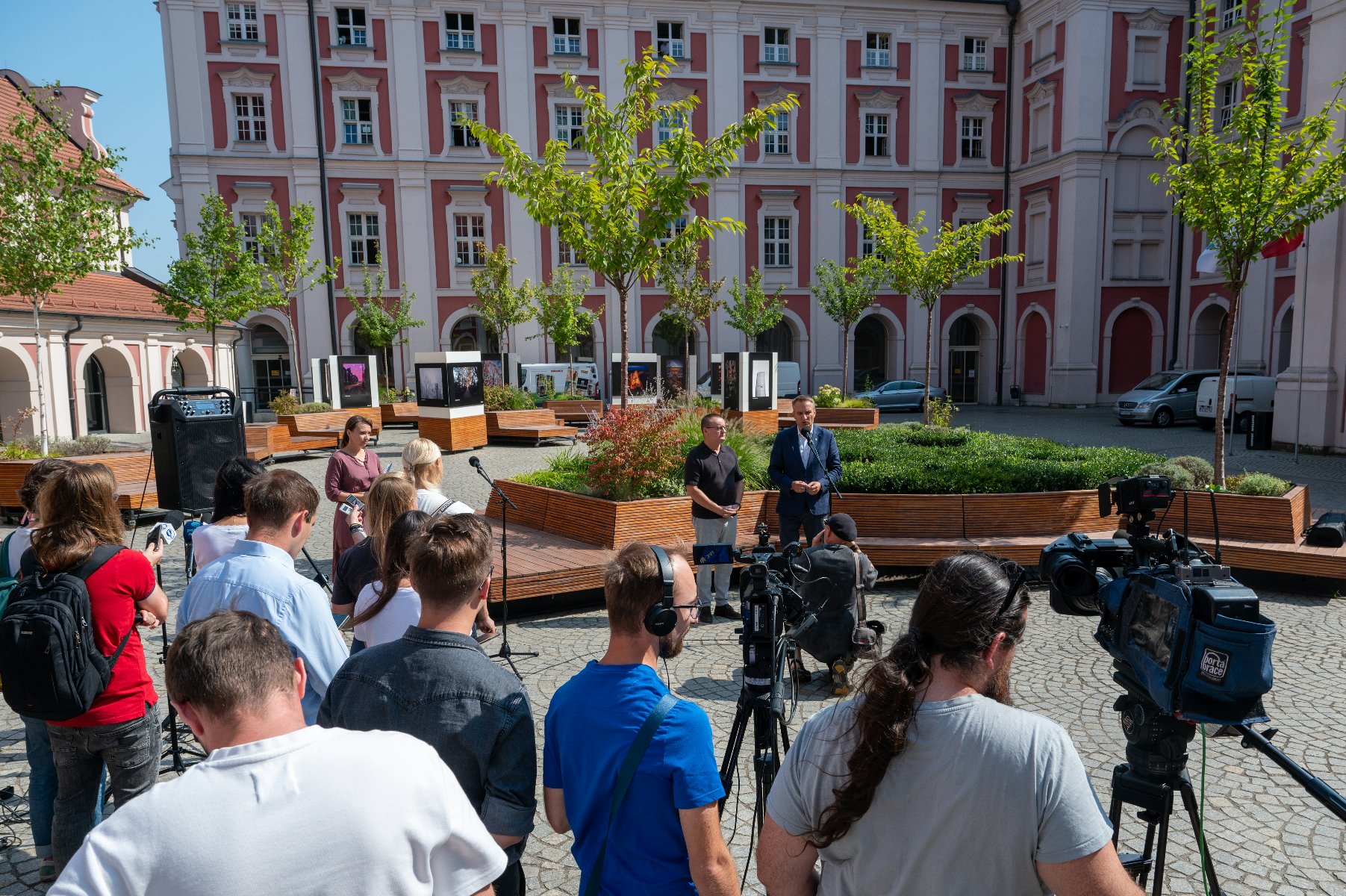 grafika przedstawia osoby zgromadzone na konferencji prasowej na dziedzińcu przez Urzędem Miasta - na pierwszym planie dziennikarze z mikforonami i kamerami, w tle mówcy - dwaj mężczyżni - grafika artykułu