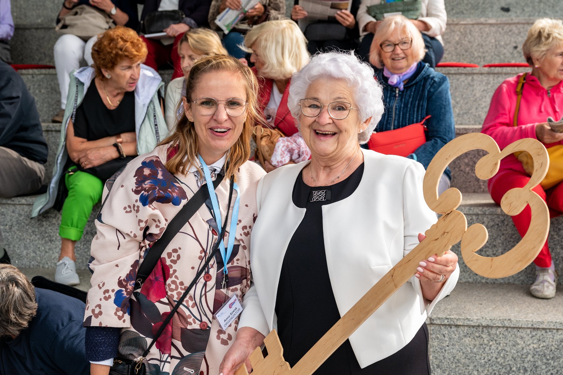 zdjęcie przedstawia starsza i młodszą kobiete, starsza trzyma w rękach duży klucz dla bram Miasta, w tle na schodach siedza seniorki - grafika artykułu