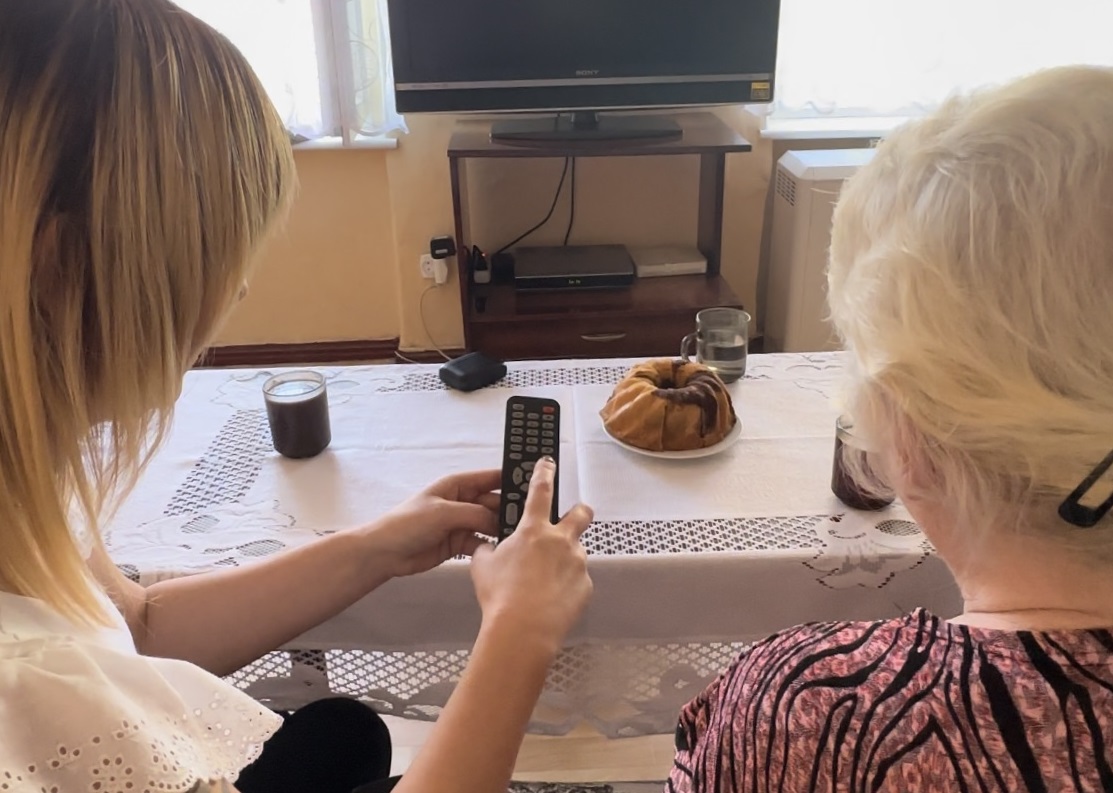 na zdjęciu dwie kobiety - młodsza pokazuje i tłumaczy starszej przyciski na pilocie do telewizora, siedza przy stole nakrytym białą serwetą. Na stole stoi babka i szklanki z kawą. Na dalszym planie widoczny jest telewizor. - grafika artykułu