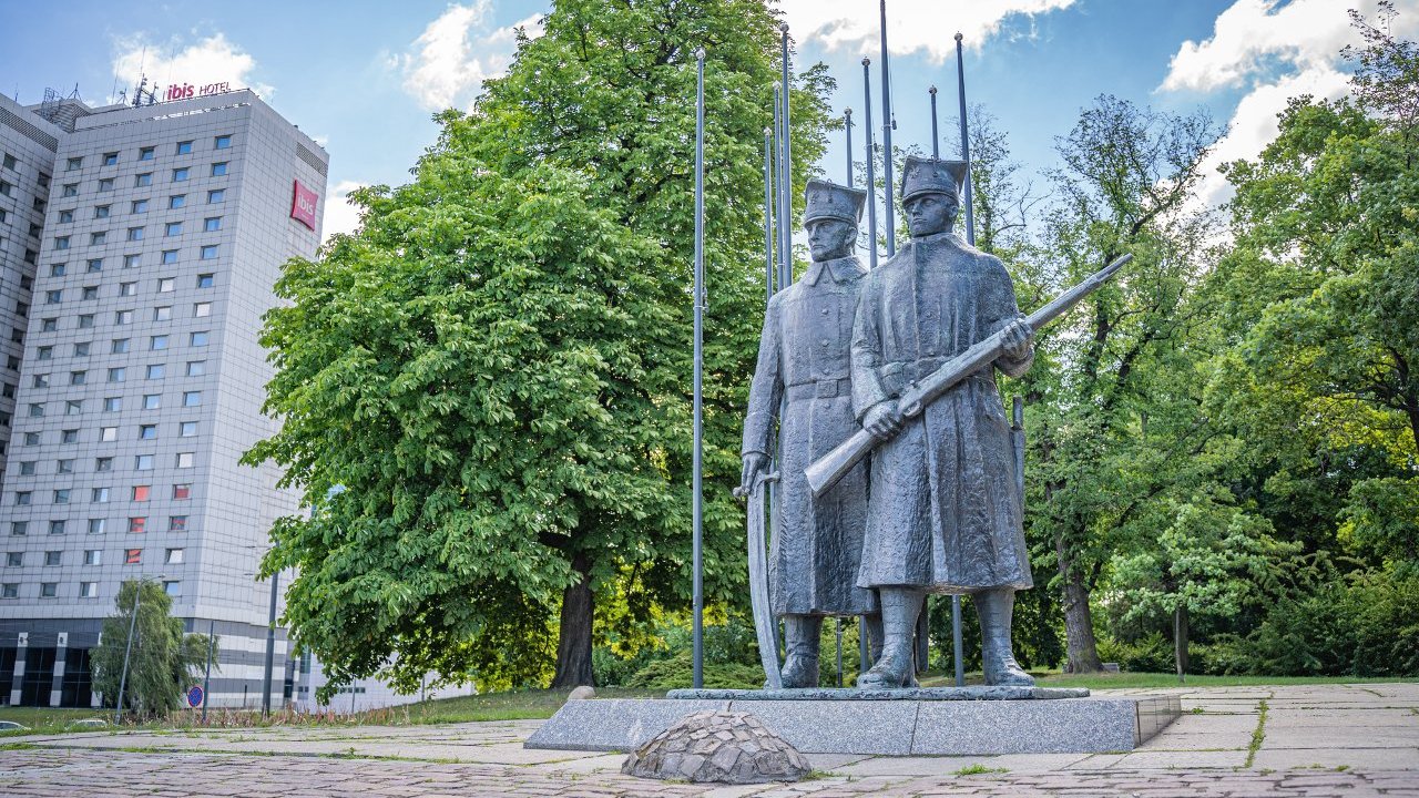 Szary pomnik dwóch żołnieży. Jeden z nich trzyma szablę, a drugi sztucer. W tle park i wieżowce.