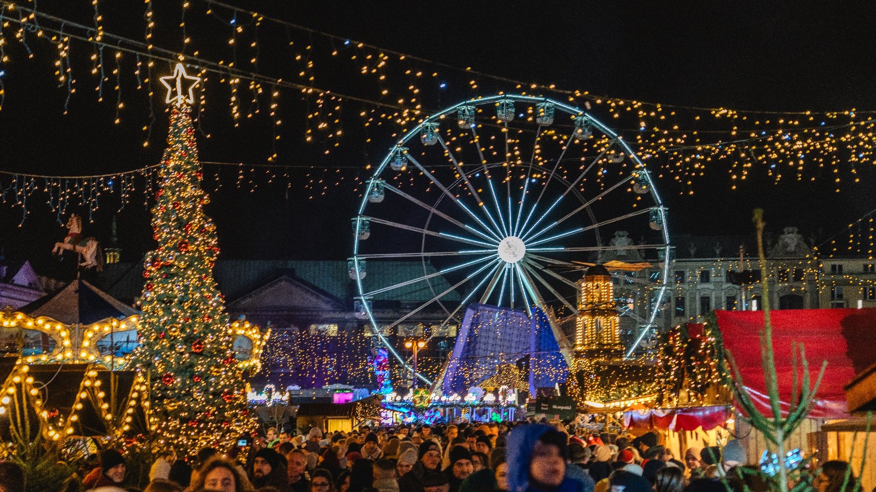 Zdjęcie przedstawia świąteczny plac Wolności. Oprócz kolorowych lampek widać na nim tłum ludzi, stoiska handlowe, choinkę i młyńskie koło.