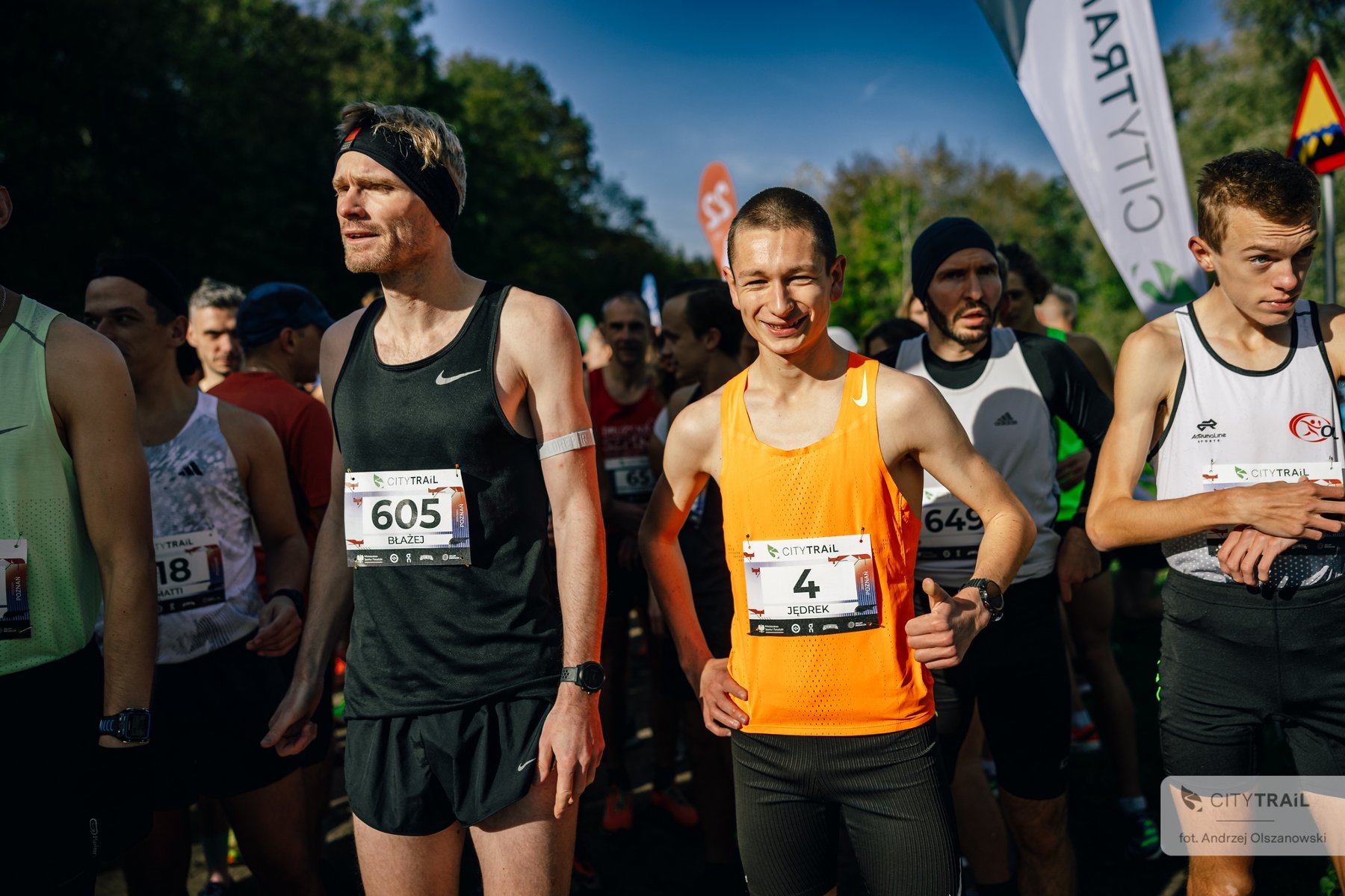 Cykl City Trail w Poznaniu, fot. Andrzej Olszanowski Grupa biegaczy stoi na starcie zawodów, czekając na rozpoczęcie biegu. Na pierwszym planie dwóch mężczyzn w sportowych koszulkach: jeden w czarnej z numerem 605, drugi w pomarańczowej z numerem 4, uśmiecha się.