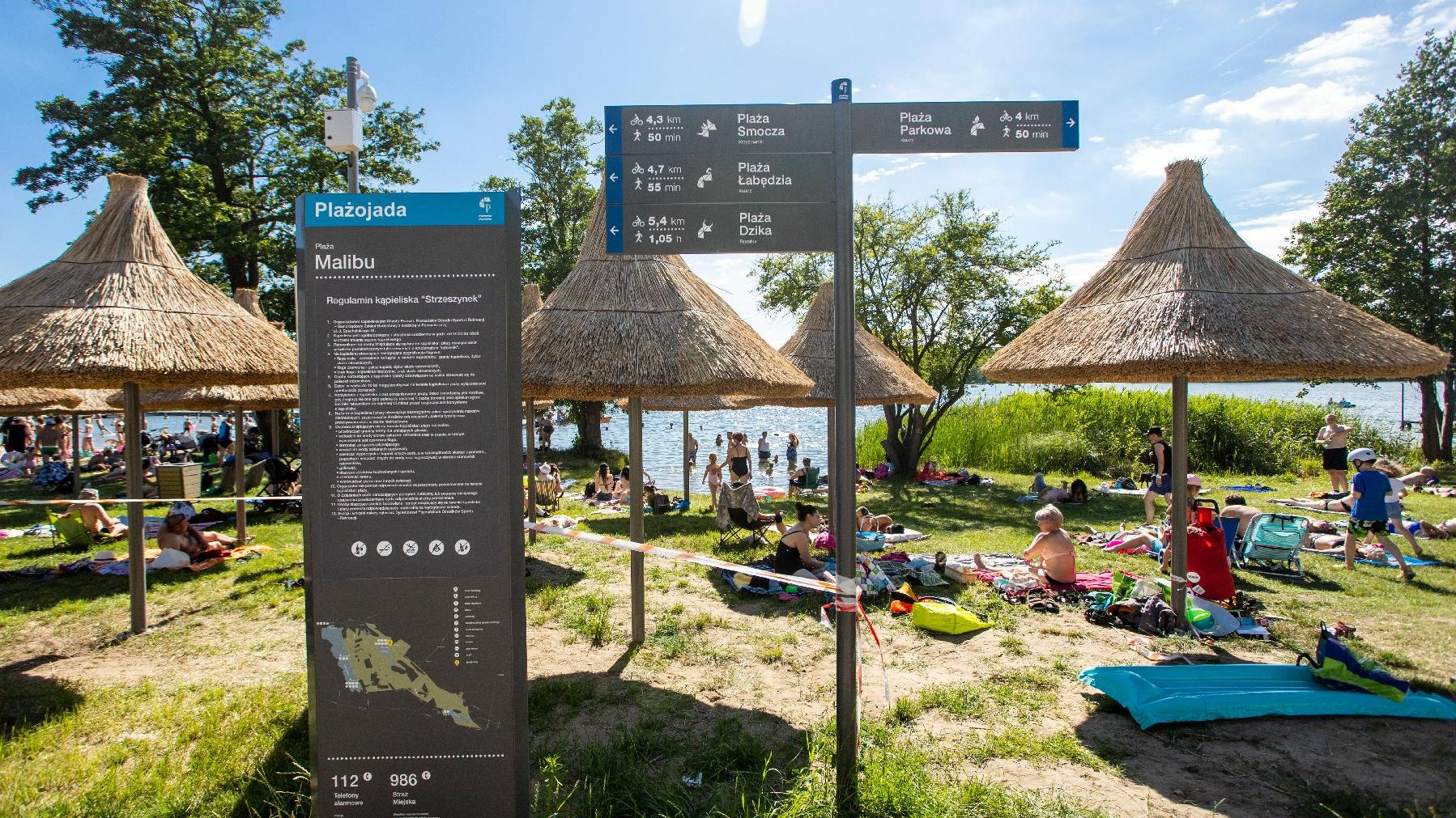 Zdjęcie przedstawia plażę - widać na nim siedzących na kocach ludzi, parasolki ze słomy oraz tablicę z opisem projektu Plażojada.