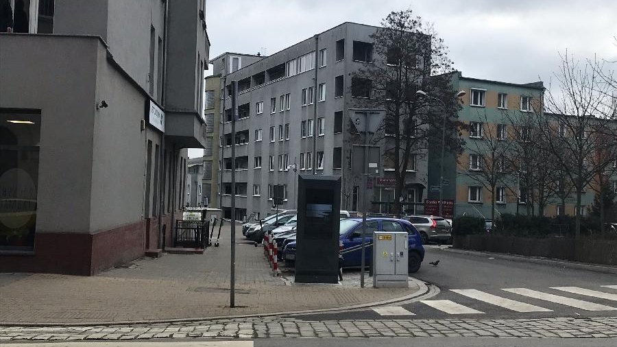 Zdjęcie przedstawia multimedialny totem, stojący na chodniku, w sąsiedztwie jezdni i zaparkowanych samochodów. W tle widać bloki.