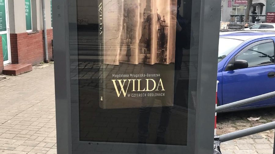 Zdjęcie przedstawia multimedialny totem. Na ekranie wyświetlona jest okładka książki pt."Wilda w czterech odsłonach". W tle widać zaparkowane samochody.