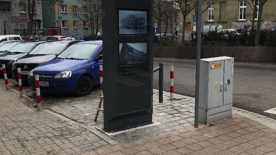 Zdjęcie przedstawia multimedialny totem. Urządzenie stoi na chodniku. Obok znajdują się zaparkowane na poboczu jezdni samochody.