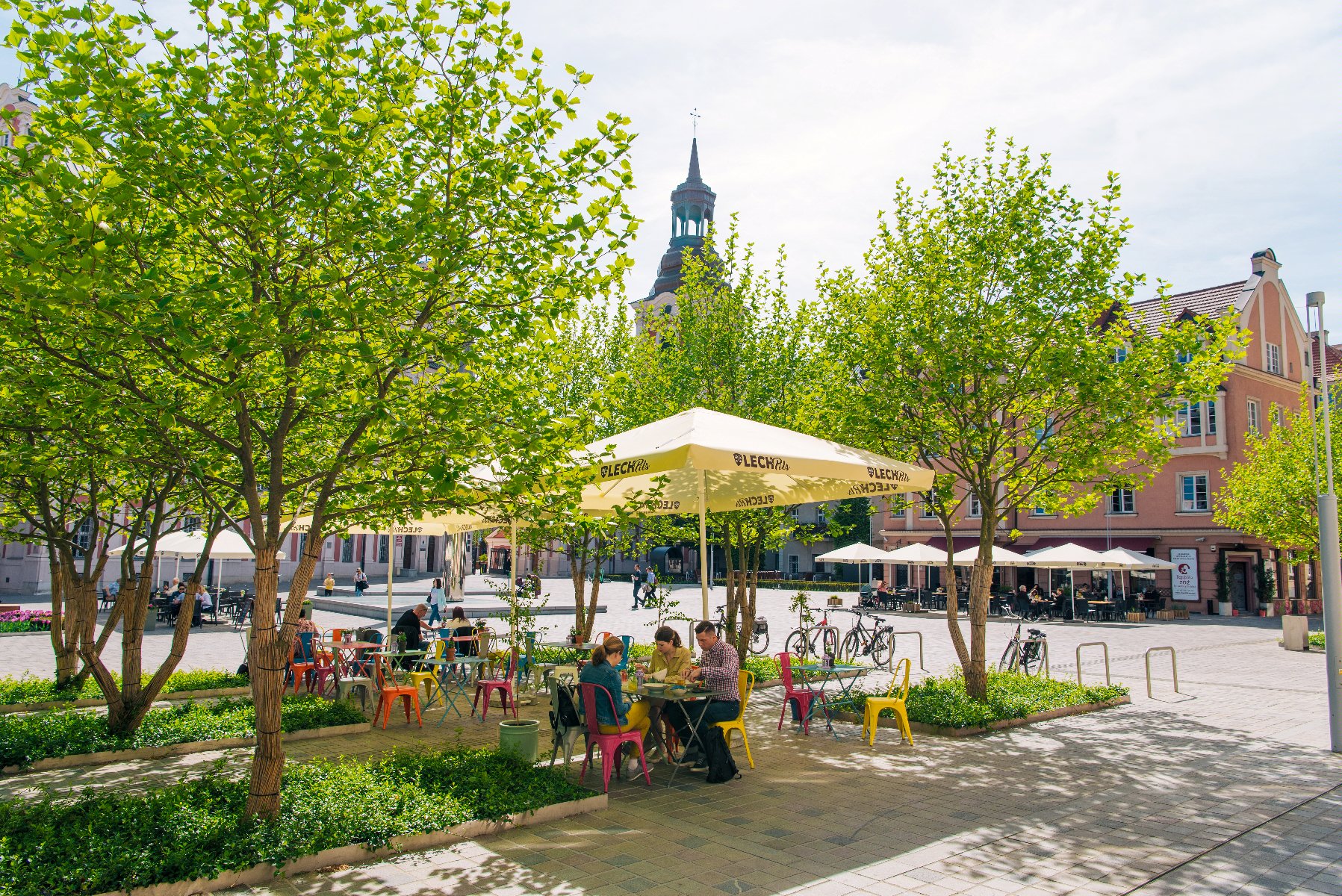 Na zdjęciu pl. Kolegiacki. Na pierwszym planie ogródek restauracyjny otoczony drzewami. W tle wieża poznańskiej Fary i kamienice. - grafika artykułu