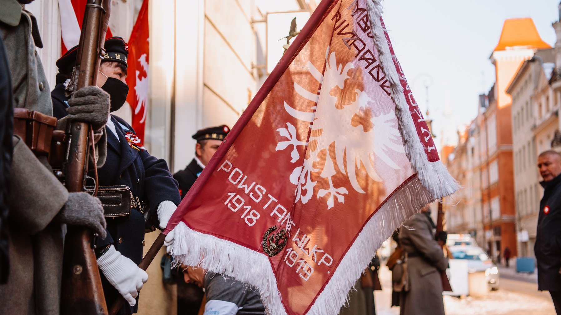 Grupa rekonstrukcyjna w mundurach z okresu Powstania Wielkopolskiego. Jedna z osób trzyma sztandar z białym orłem oraz datą 1918-1919.