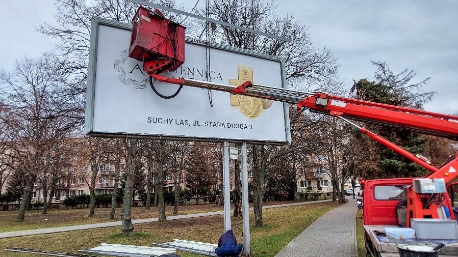 Na zdjęciu widać wielkoformatową reklamę, obok na chodniku stoi ciężarówka z wysięgnikiem.