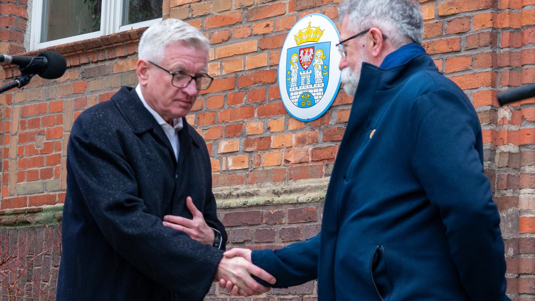 Na zdjęciu dwóch mężczyzn podających sobie ręce, jednym z nich jest prezydent Poznania, za nimi ceglana ściana, na niej herb Miasta