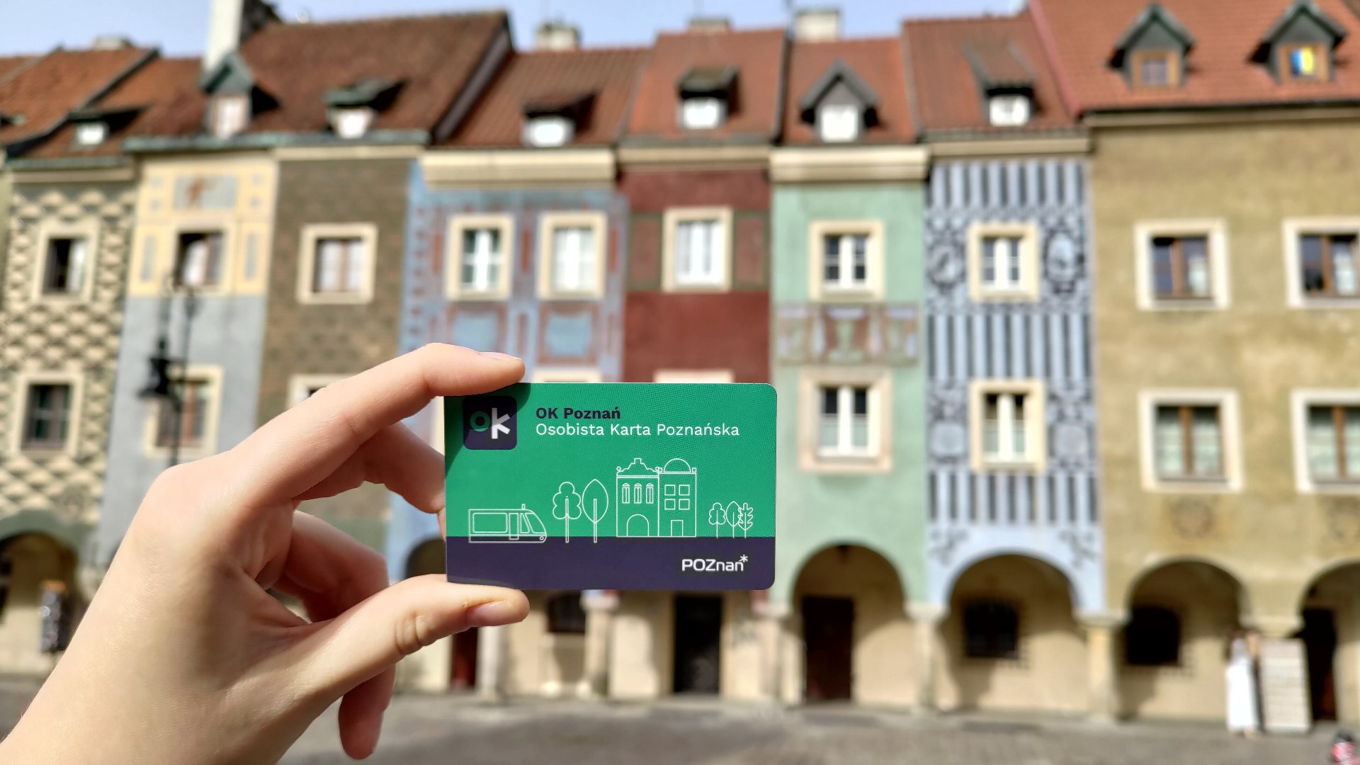 Na zdjęciu dłoń trzymająca kartę ok Poznań na tle domków budniczych - grafika artykułu