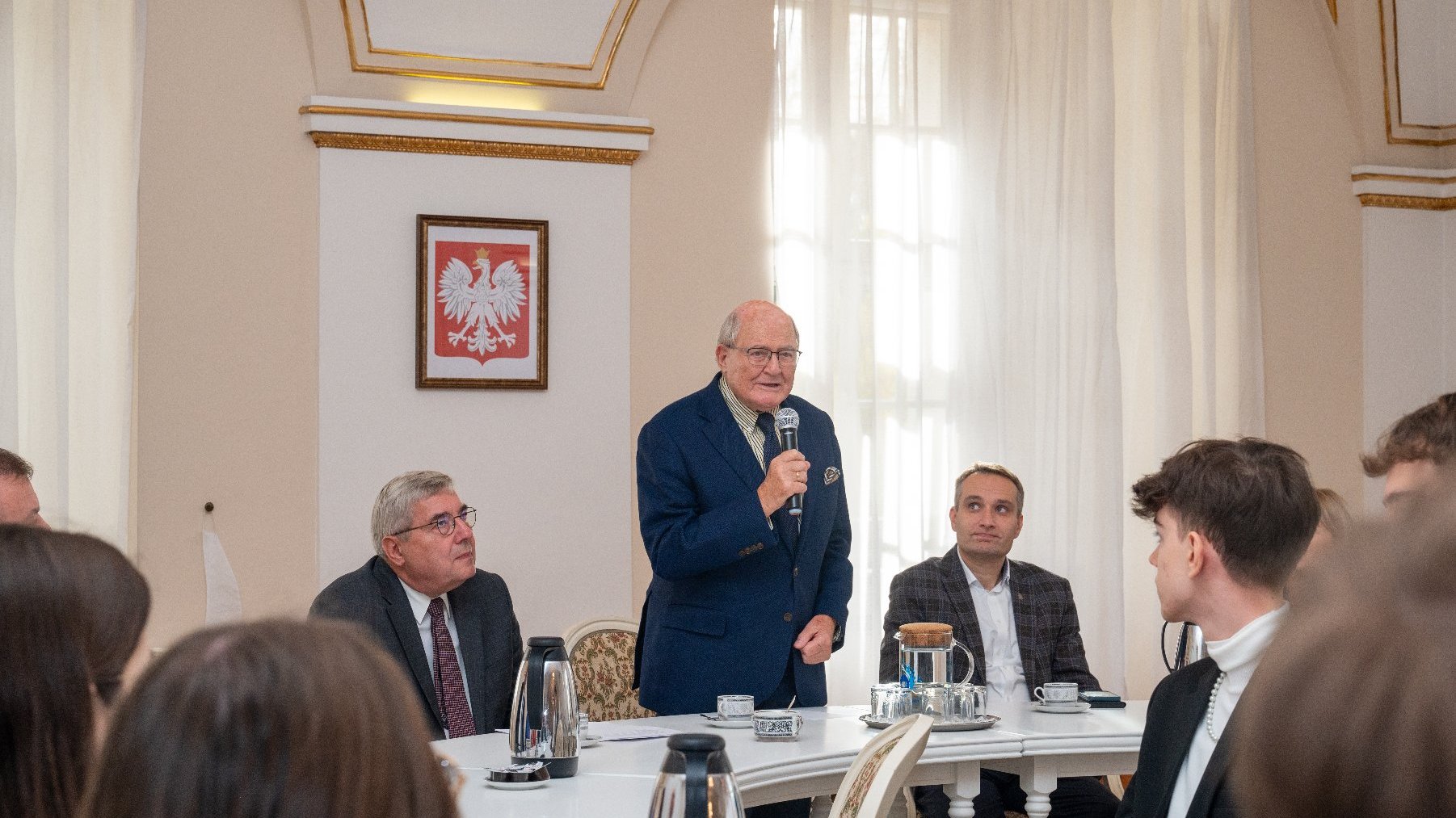 Zdjęcie przedstawia ludzi siedzących przy stole oraz stojącego mężczyznę, trzymającego w ręku mikrofon.