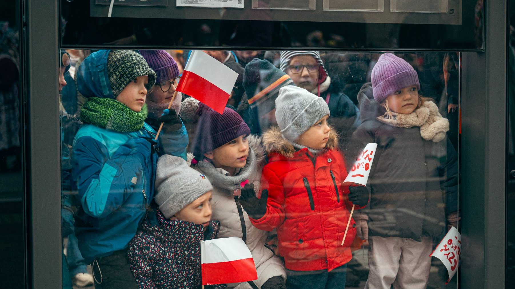 Dzieci obserwujące przez szybę przystanku. Niektóre trzymają biało-czerwone flagi