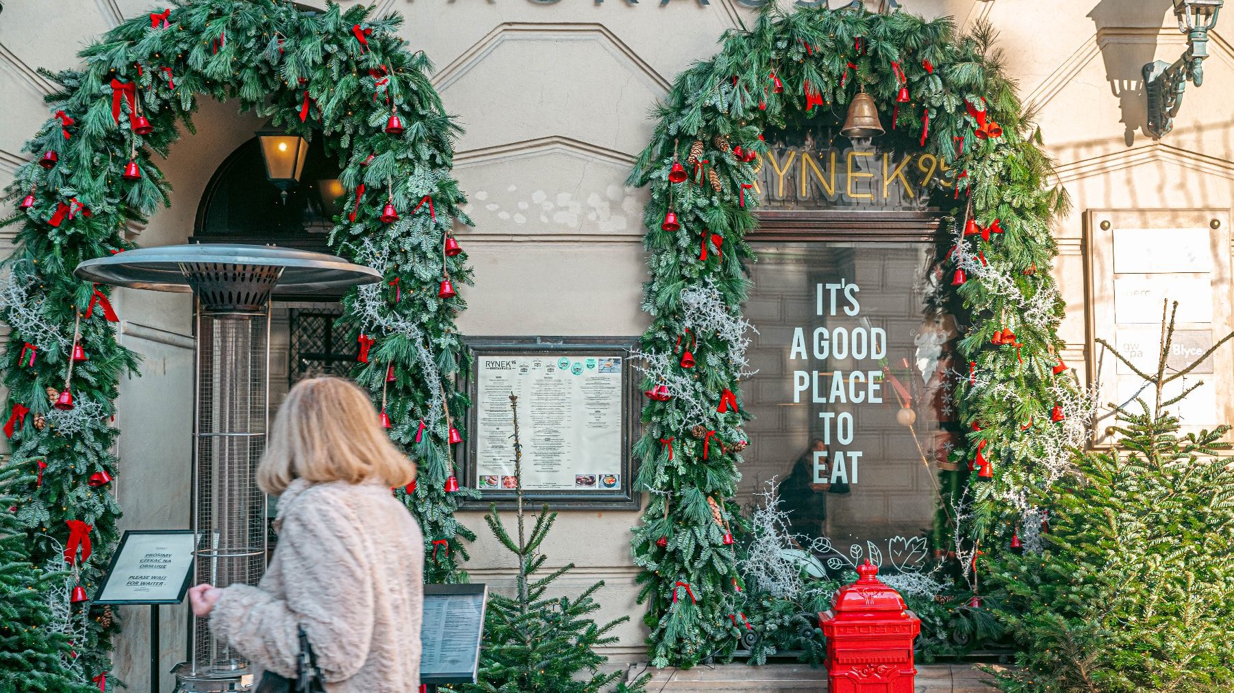 "Wejście do restauracji ozdobione świątecznymi girlandami i czerwonymi wstążkami, prawdopodobnie na sezon bożonarodzeniowy. Na drzwiach widnieje napis: "IT'S A GOOD PLACE TO EAT." Przed wejściem przechodzi osoba z jasnymi włosami i w jasnym płaszczu. Przed restauracją znajduje się czerwona skrzynka pocztowa. Na górze obrazu widoczny jest napis "RESTAURACJA", co wskazuje, że to wejście do restauracji. Obok wejścia znajduje się menu w szklanej gablocie.