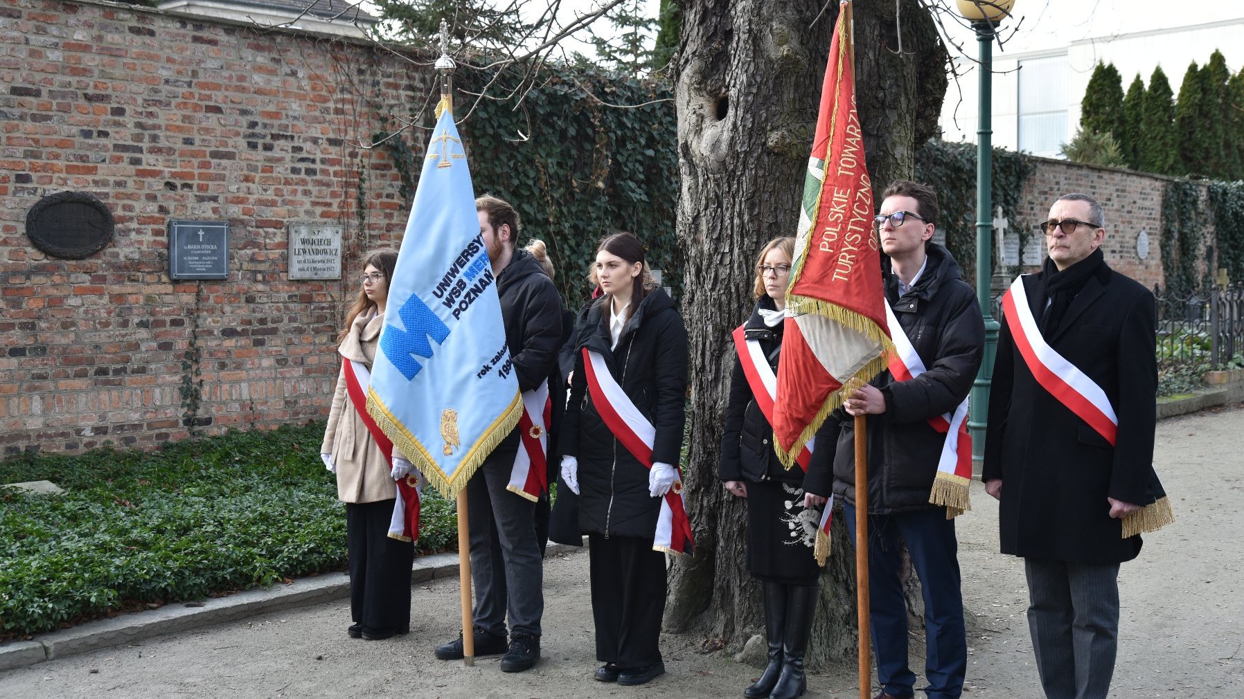 Grupa osób stoi przed ceglaną ścianą z tablicami pamiątkowymi. Trzymają flagi i noszą szarfy, co sugeruje formalne lub ceremonialne wydarzenie. Jedna z flag jest niebieska, a druga czerwona. Osoby są ubrani formalnie, co wskazuje na uroczysty lub oficjalny charakter zgromadzenia. Miejsce wydarzenia znajduje się na zewnątrz, prawdopodobnie w miejscu historycznym lub pamięciowym.