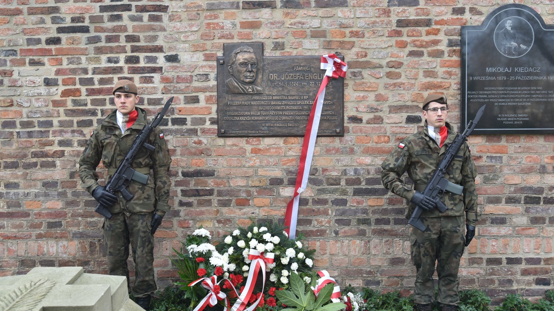 Obraz przedstawia dwóch żołnierzy w mundurach wojskowych stojących na straży przed ceglanym murem z dwiema tablicami pamiątkowymi. Żołnierze trzymają karabiny i są ustawieni po obu stronach ekspozycji pamięciowej, która obejmuje wieniec z czerwono-białymi wstążkami i kwiatami. Tablice na ścianie wydają się być pamiątkowe, z jedną poświęconą dr Józefowi Englichowi, a drugą Mikołajowi Kiedaczowi.