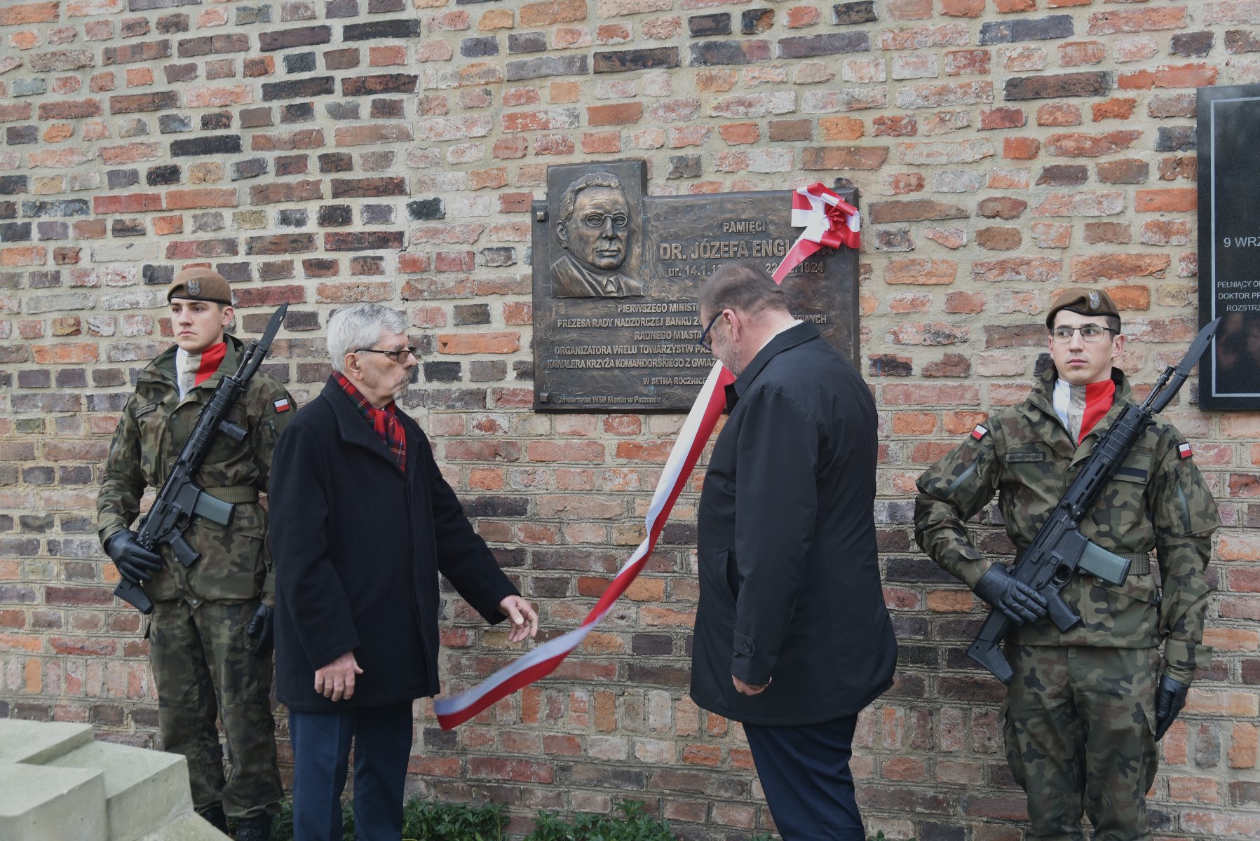 Zdjęcie przedstawia ceremonię, odbywającą się przed ceglaną ścianą z umieszczoną na niej tablicą pamiątkową. Obecnych jest czterech uczestników: dwóch w mundurach wojskowych trzymających karabiny oraz dwóch ubranych w cywilne ubrania. Dwaj cywile zdają się odsłaniać lub prezentować tablicę, ponieważ trzymają wstążkę przymocowaną do niej. Tablica poświęcona jest dr. Józefowi Englichowi. - grafika artykułu