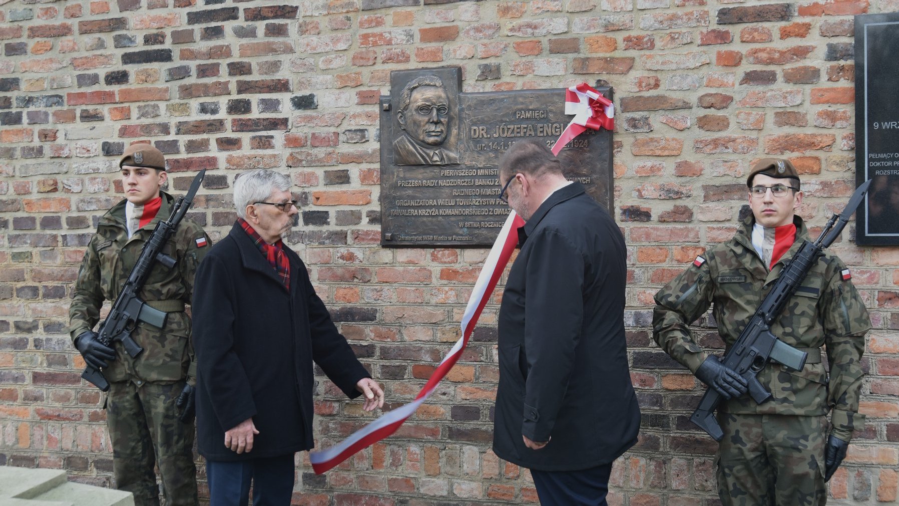 Zdjęcie przedstawia ceremonię, odbywającą się przed ceglaną ścianą z umieszczoną na niej tablicą pamiątkową. Obecnych jest czterech uczestników: dwóch w mundurach wojskowych trzymających karabiny oraz dwóch ubranych w cywilne ubrania. Dwaj cywile zdają się odsłaniać lub prezentować tablicę, ponieważ trzymają wstążkę przymocowaną do niej. Tablica poświęcona jest dr. Józefowi Englichowi.