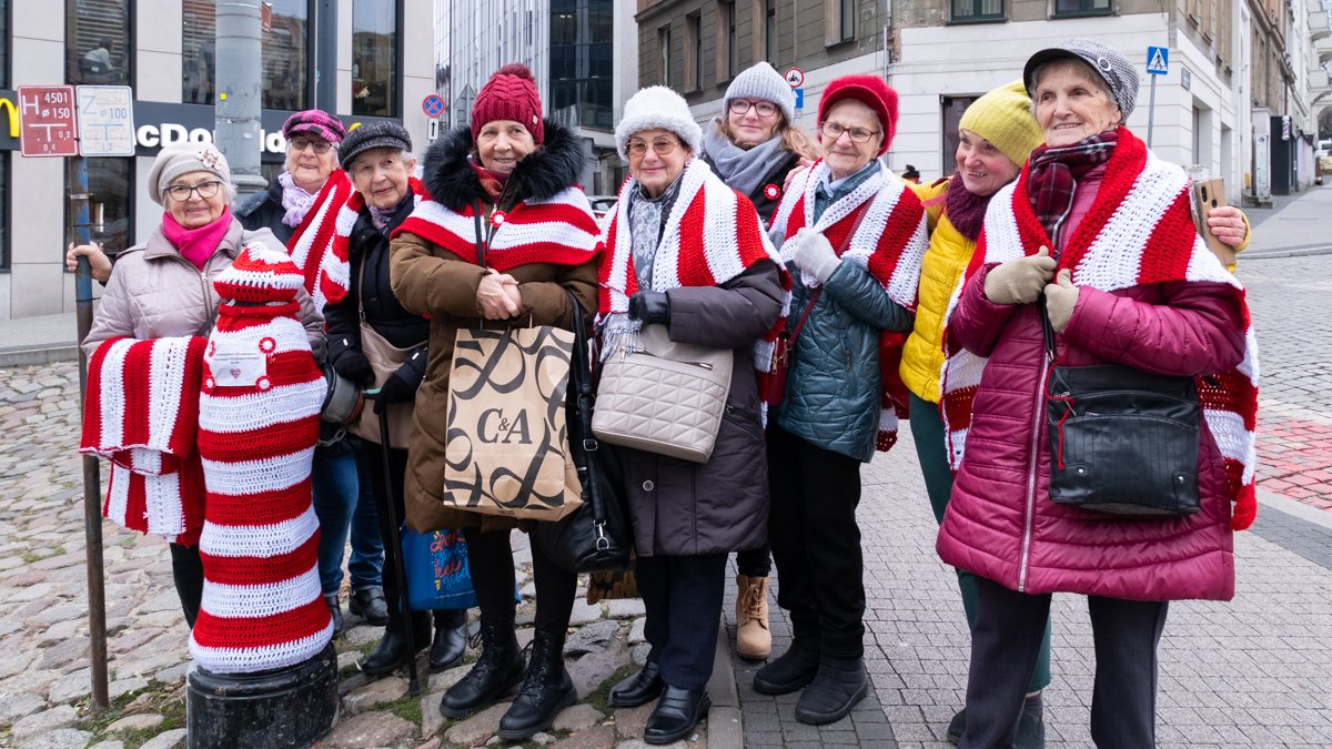 Zdjęcie przedstawia grupę starszych kobiet pozujących do wspólnego zdjęcia na ulicy. Wszystkie mają zarzucone na plecach biało czerwone kubraczki.