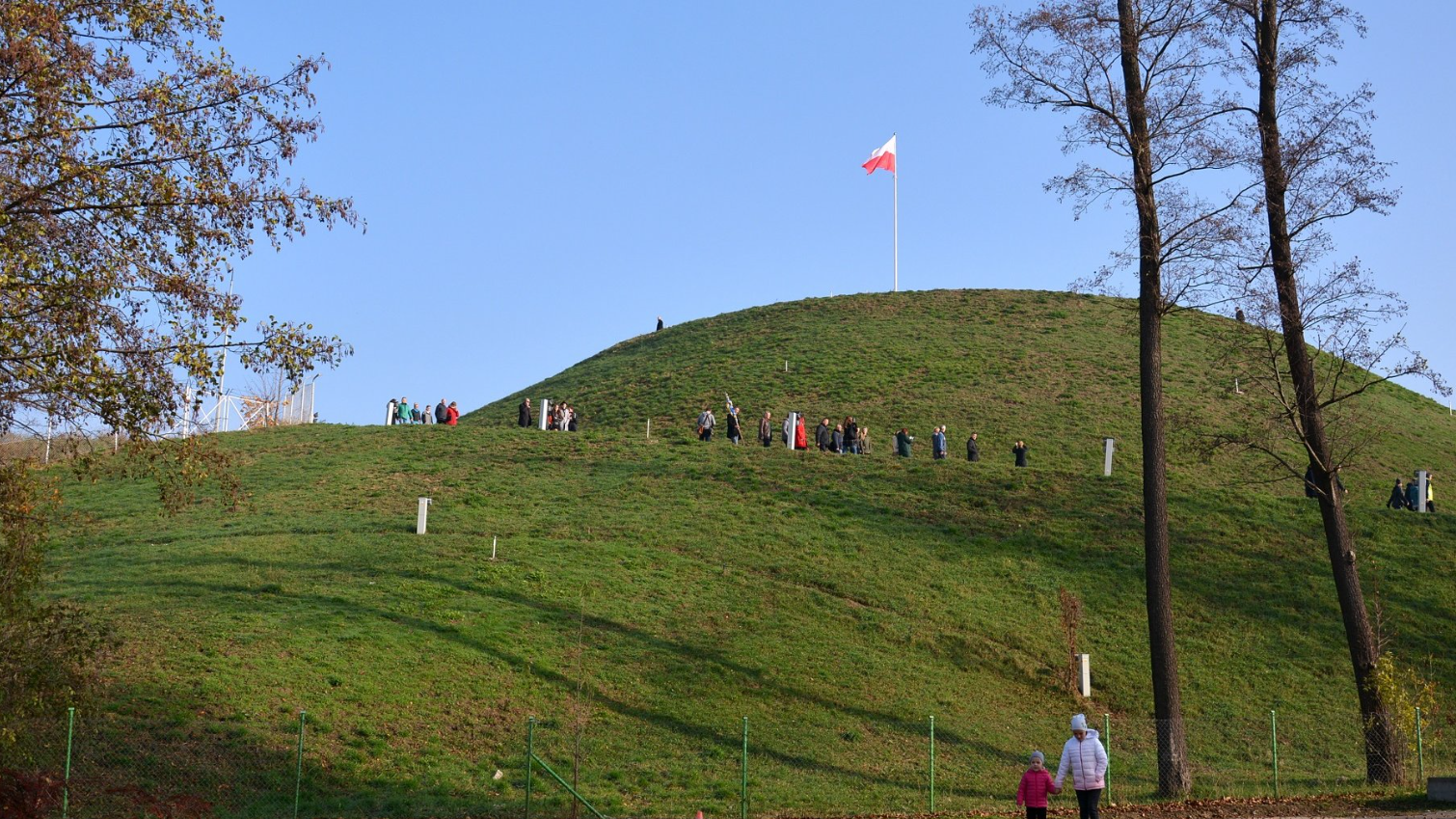 "Zdjęcie przedstawia wzgórze pokryte trawą z masztem flagowym na szczycie, na którym powiewa czerwono-biała flaga. Kilka osób wspina się na wzgórze, tworząc linię od dołu do góry. Wzgórze jest otoczone ogrodzeniem, a po prawej stronie obrazu widać kilka drzew. Na niebie jest jasno i bezchmurnie, co wskazuje na słoneczny dzień - grafika artykułu