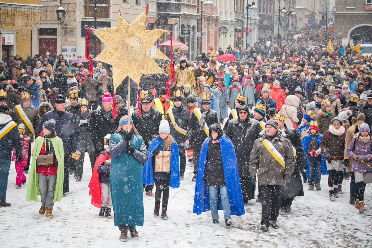 Zdjęcie przedstawia tłum ludzi idących przez Stary Rynek. Osoby w pierwszym rzędzie mają przebrania i niosą dużą złotą gwiazdę. Pada śnieg. - grafika artykułu