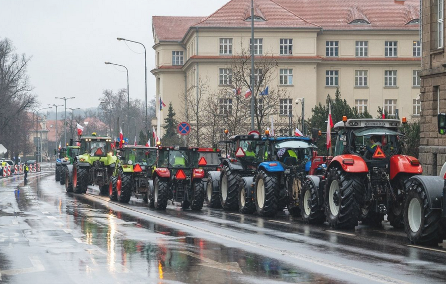 Kilka traktorów ustawionych jeden za drugim na ulicy, asfalt mokry od deszczu, za traktorami budynki - grafika artykułu