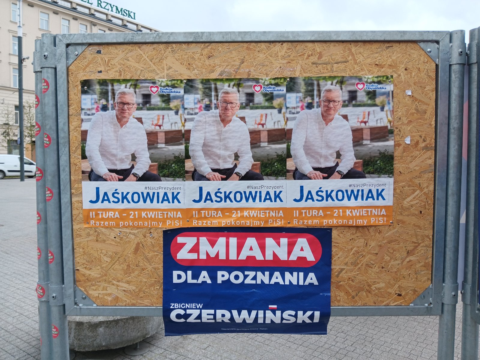 Tablica z plakatami wyborczymi, na górze tablicy trzy plakaty przedstawuająca mężczyznę w białej koszuli, siwych włosach i okularach, który siedzi na ławce, za nim drzwa, podpis "Jaśkowiak", na plakacie na dole napis "Zmiana dla Poznania Czerwiński" - grafika artykułu