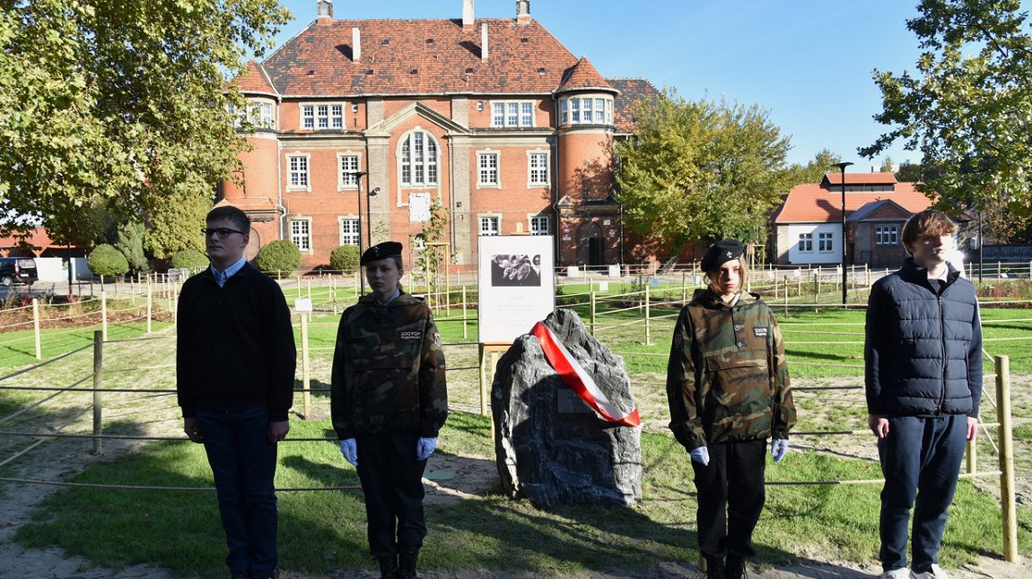 harcerze i uczniowie, cztery osoby, pełnią wartę przed pamiątkowym głazem z tablicą, głaz przepasany jest biało czerwoną wstęgą, za nimi budybnek szkoły z czewonej cegły, ze spadzistym dachem