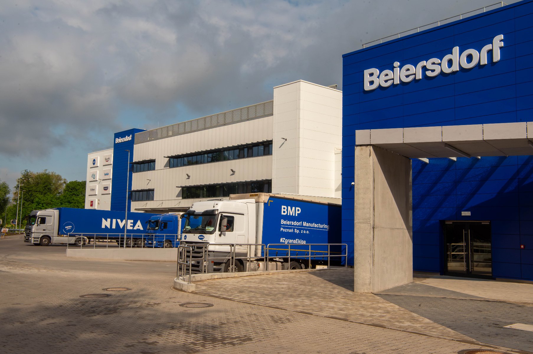 W Poznaniu otwarto rozbudowane Centrum Produkcyjne Beiersdorf Zewnętrzny widok na budynki fabryki Beiersdorf w Poznaniu, z kilkoma ciężarówkami stojącymi na placu załadowczym. Na pierwszym planie niebieski budynek z logo "Beiersdorf".