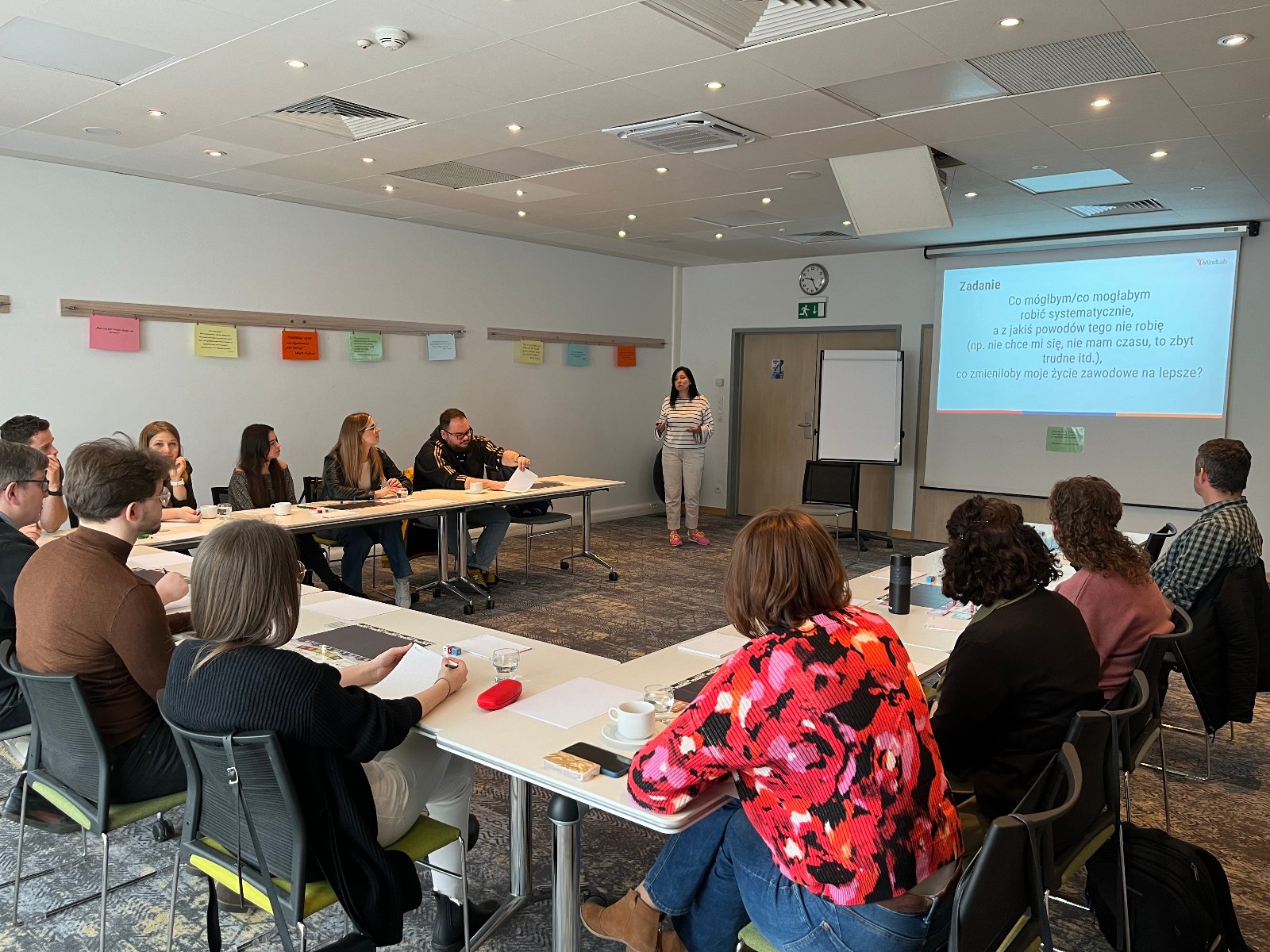 A group of people in the middle of a workshop. They are sitting at tables arranged in a U-shape. A task is displayed on a projector. - grafika artykułu