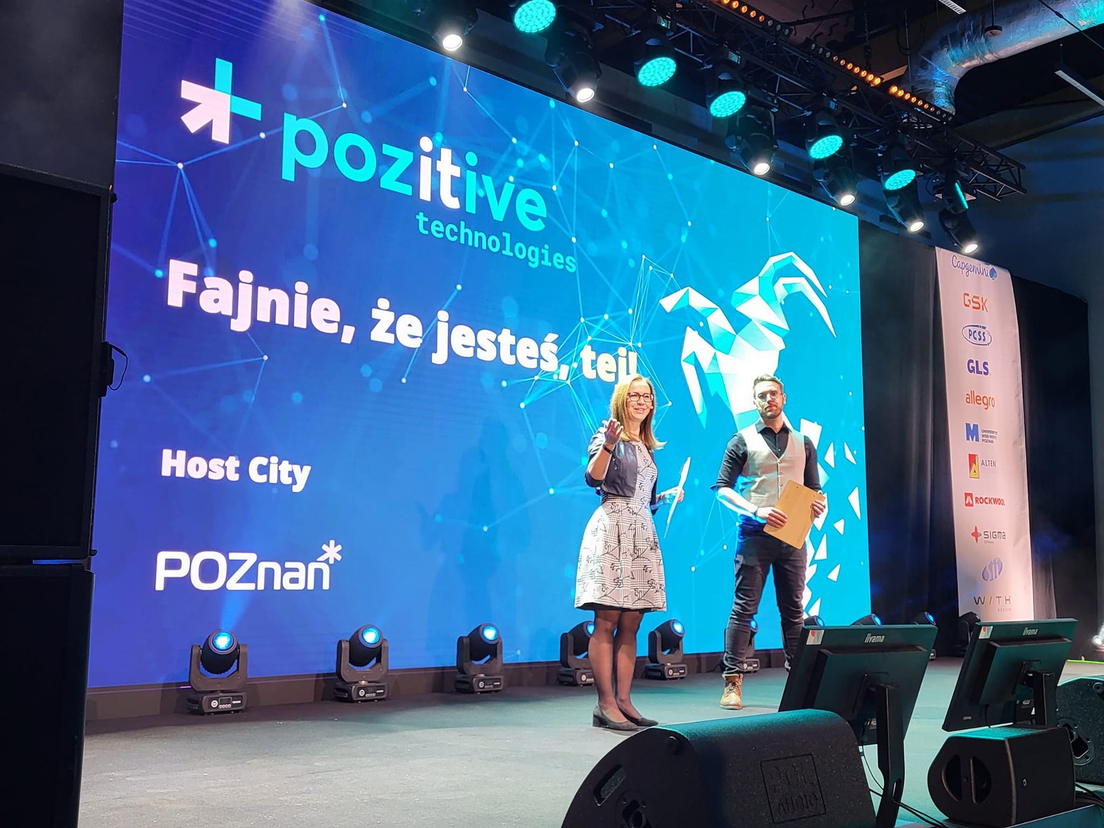 The photo shows 2 people, a woman and a man - the convenor of Pozitive Technoologies 2023. They are standing in front of a screen with the conference logo and the caption "Cool that you are, this one!'. You can also see Host City - Poznan - grafika artykułu