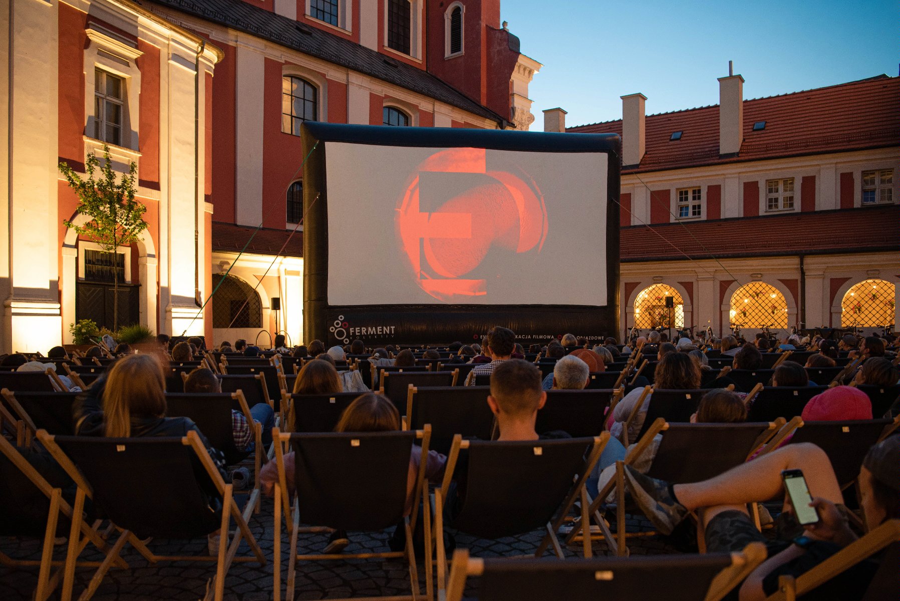 Zdjęcie przedstawia ludzi na leżakach oglądających film na telebimie na dziedzińcu urzędu miasta. - grafika artykułu