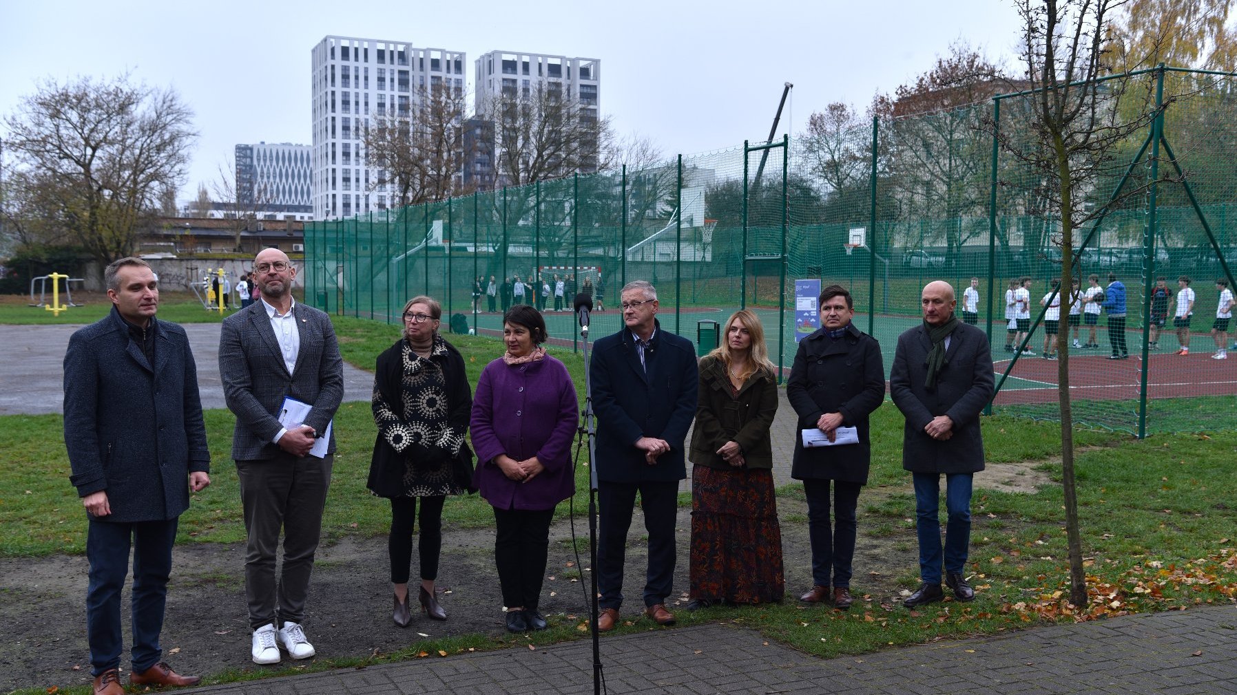 Zdjęcie przedstawia uczestników konferencji prasowej. Za ich plecami znajduje się boisko.