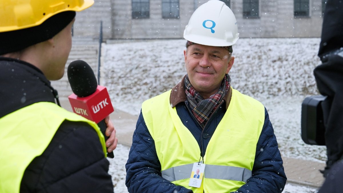 Zdjęcie przedstawia dwóch mężczyzn. Jeden stoi tyłem i trzyma w ręku mikrofon, drugi - w odblaskowej kamizelce i kasku - stoi przodem do aparatu.