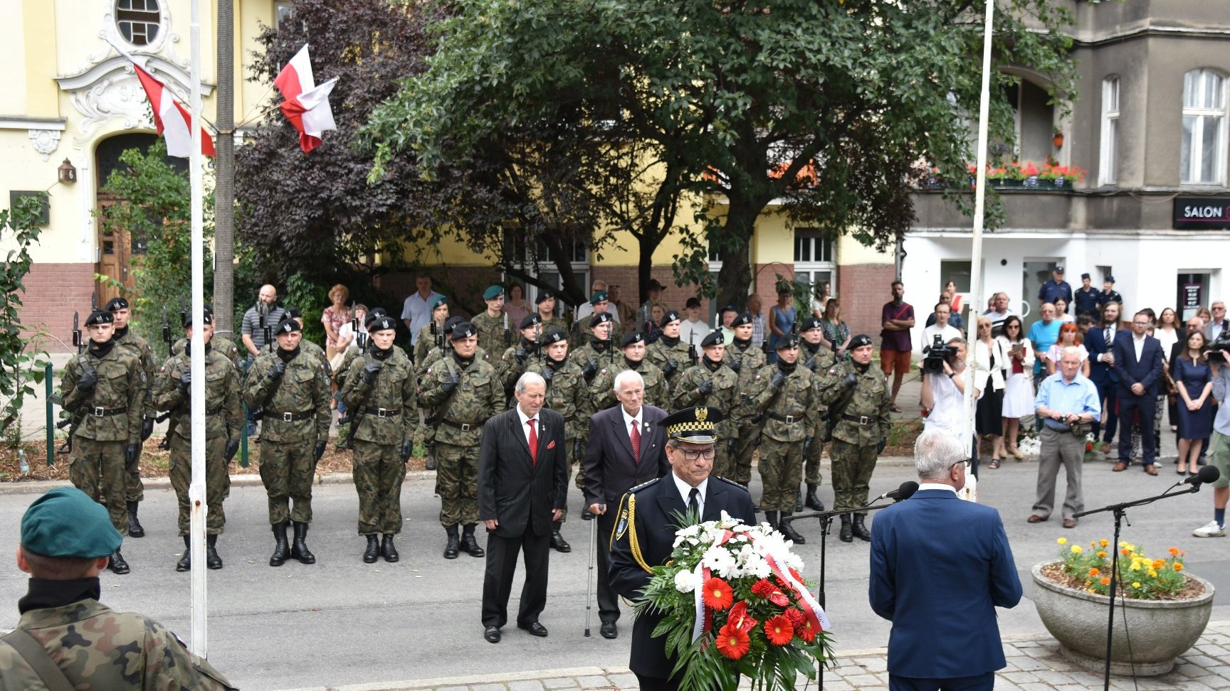 Na zdjeciu dwóch mężczyzn składających kwiaty, za nimi żołnierze