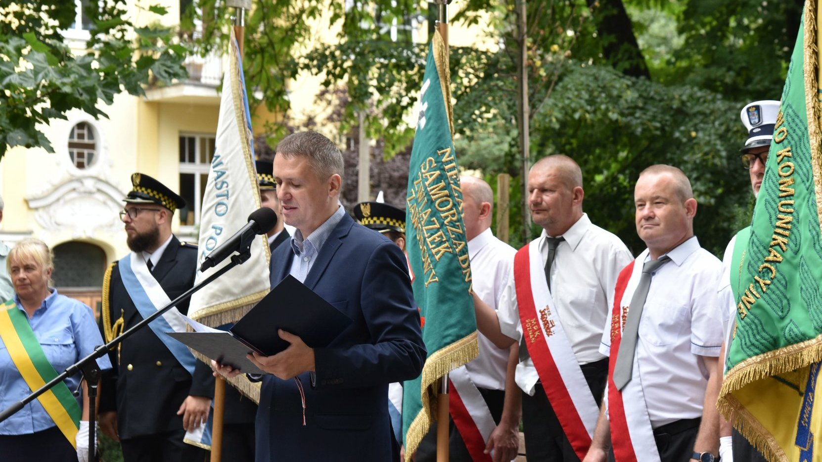 Na zdjęciu mężczyzna mówiący coś przez mikrofon, za nim poczty sztandarowe