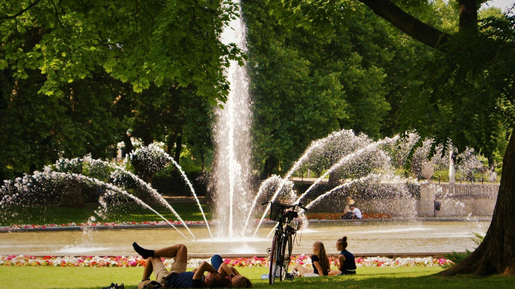 Zdjęcie przedstawia ludzi leżacych na trawie. Za ich plecami znajduje się fontanna.