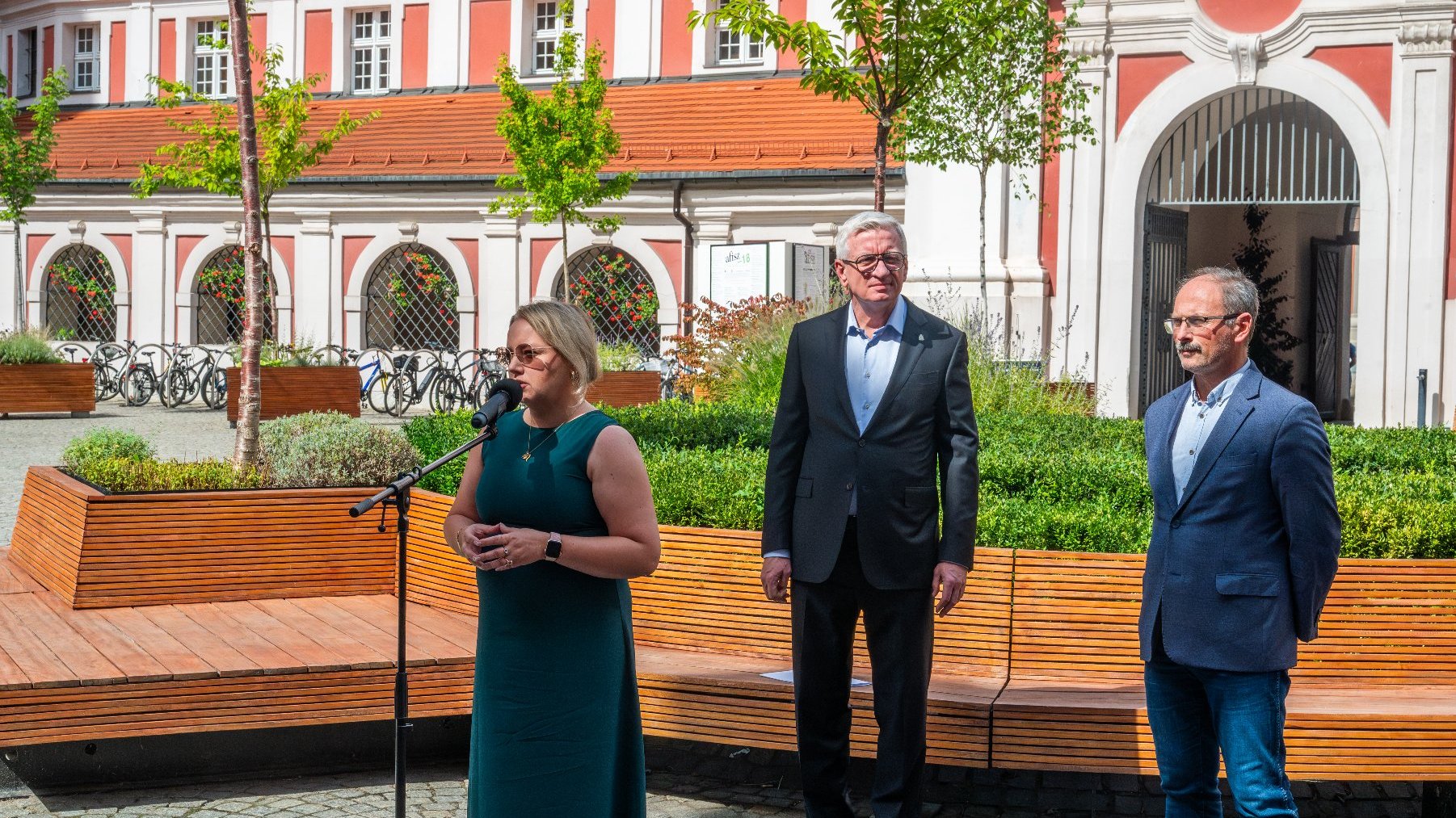 Na zdjęciu trzy osoby podczas briefingu prasowego, na pierwszym planie kobieta, za nią dwaj mężczyźni