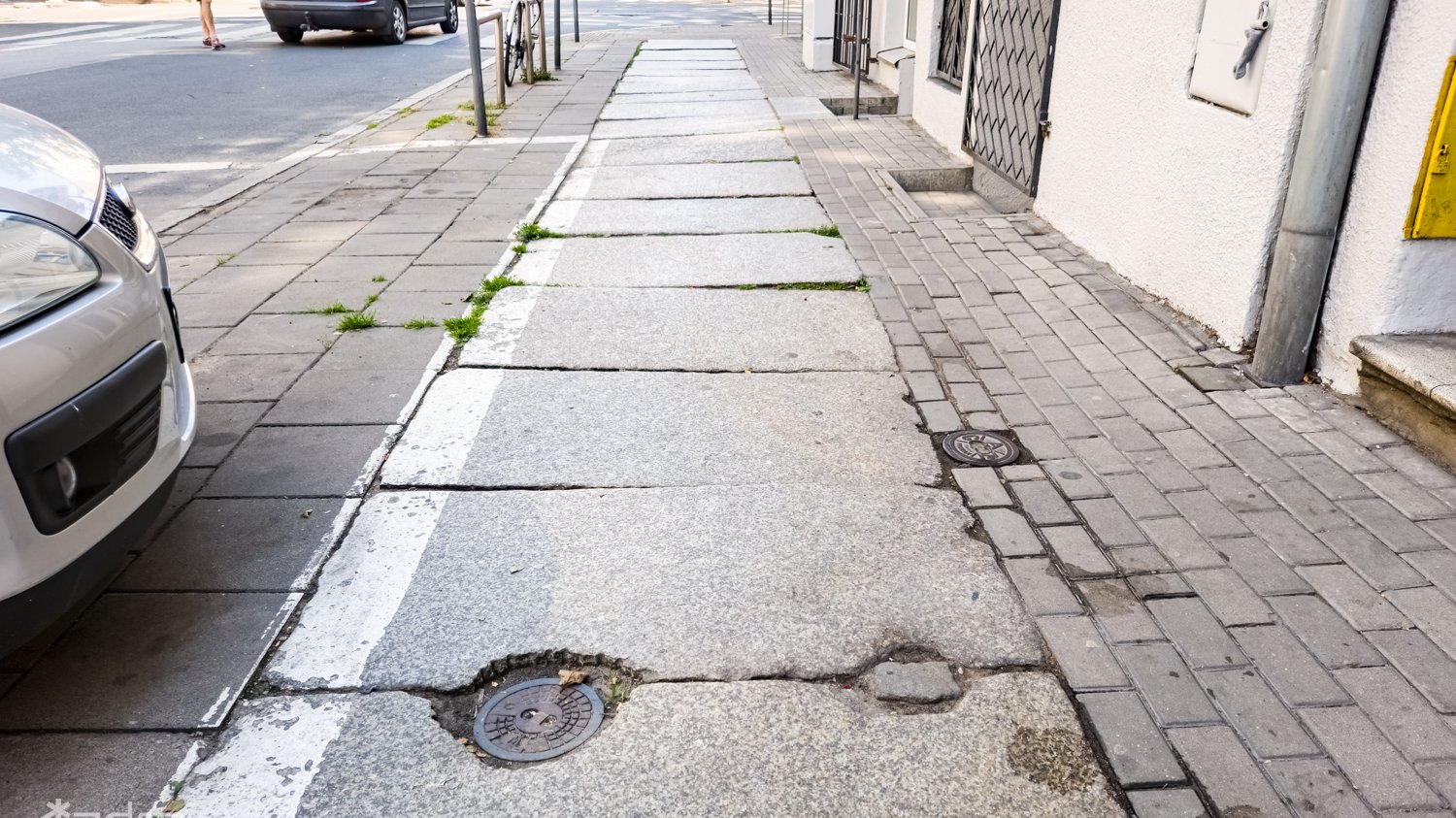 Widać zniszczony chodnik i parkujące samochody