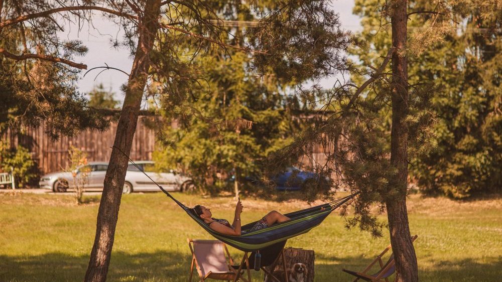 Kobieta leży w hamaku czytając coś w telefonie. Dookoła soczysta zieleń, z tyłu zaparkowane samochody.