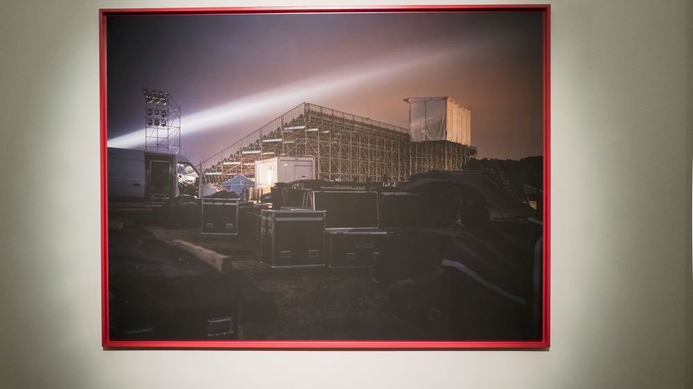 Fotografia w czerwonej ramce na białej ścianie. Przedstawia przygotowania do przedstawienia plenerowego - rozstawiona trybuna dla widzów, za nią stoi sprzęt do nagłośnienia.